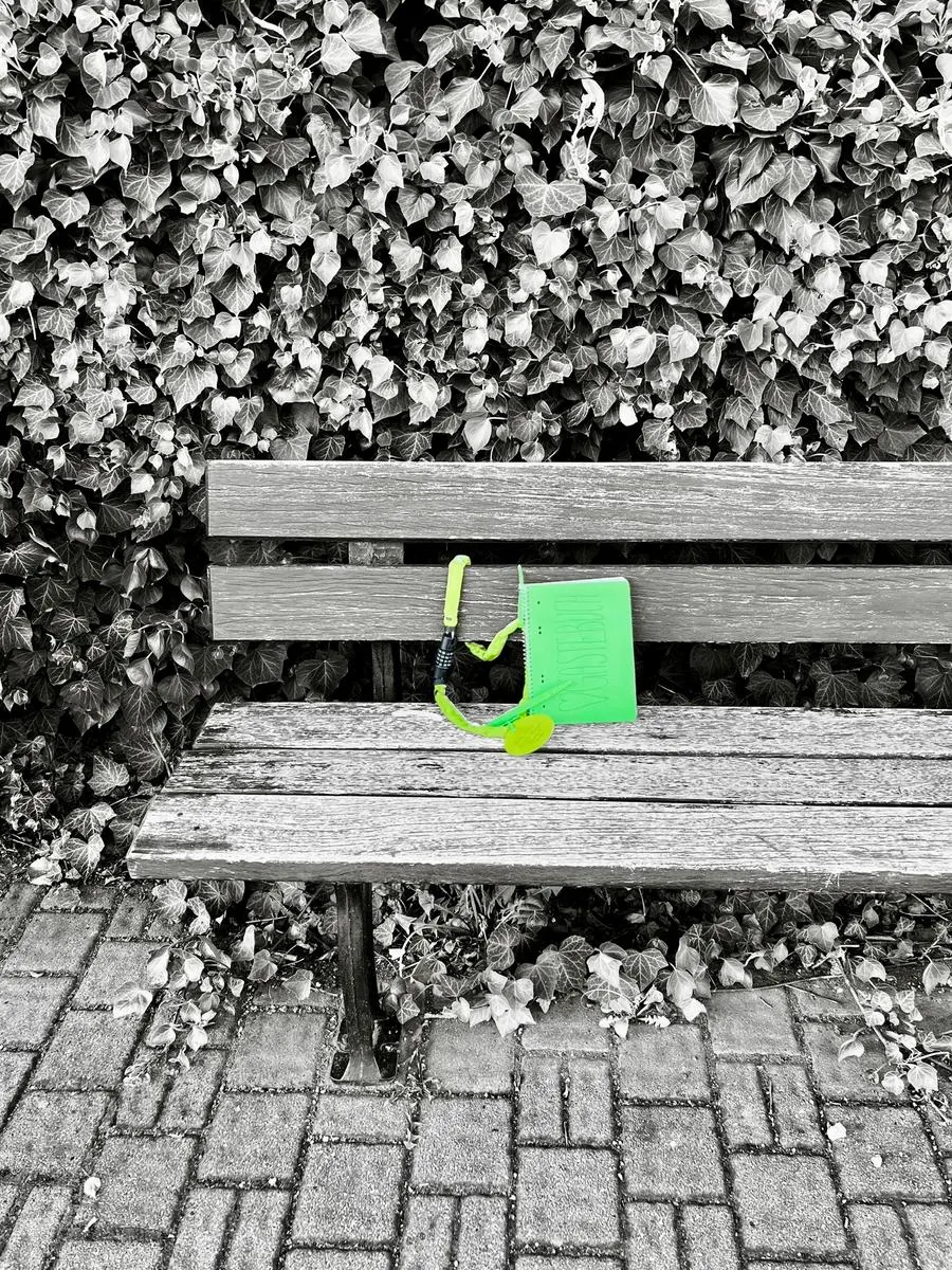 Schwarz-weiß-Foto einer Parkbank vor einer Efeuhecke, darauf farbig hervorgehoben das grüne Gästebuch mit Kette.