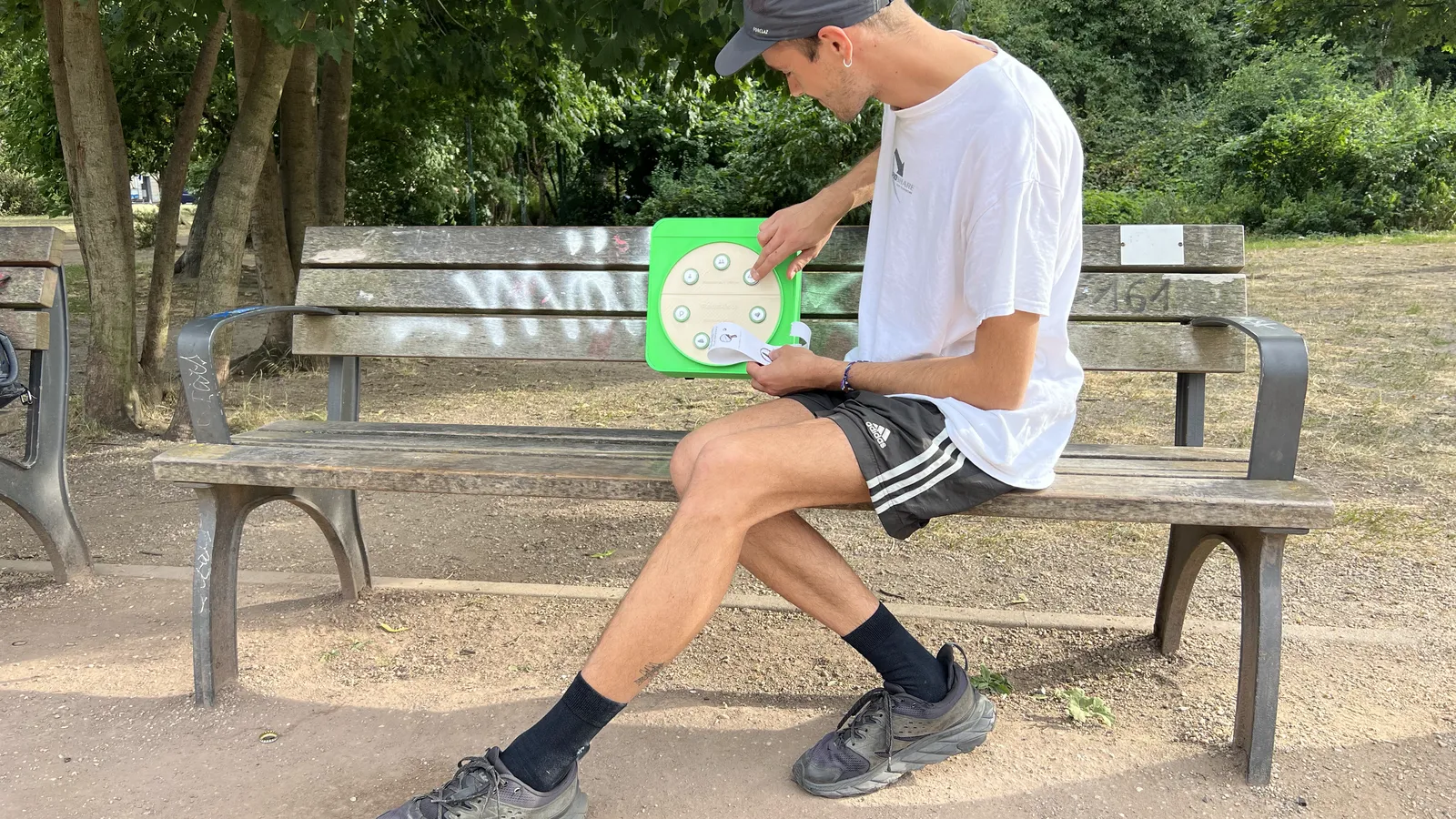 Ein Mann sitzt auf einer Parkbank und liest einen Zettel, den er gerade aus der grünen Redekiste an der Rückenlehne gezogen hat.