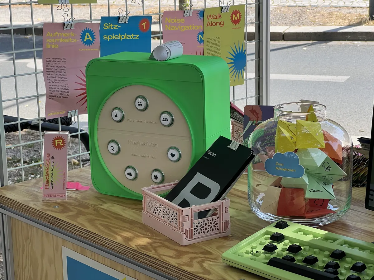 Die grüne Redekiste auf einem Holztisch, daneben ein Glas mit bunten Papierfiguren und Plakate im Hintergrund.